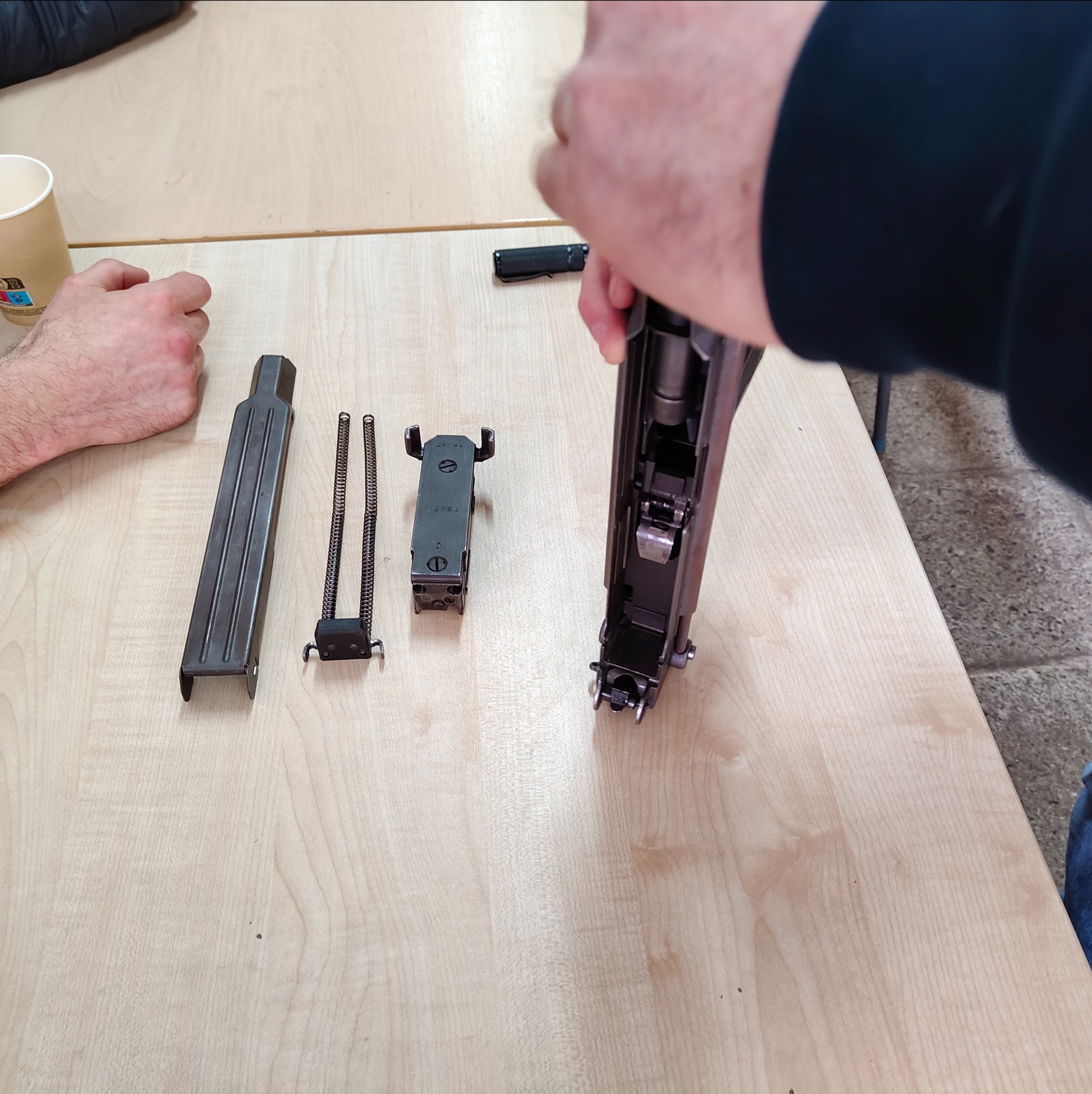 Rozłożony karabin na stole podczas szkolenia z obsługi broni. Widoczne elementy: magazynek, sprężyny, zamek i korpus. Ręce instruktora w trakcie demonstracji.
