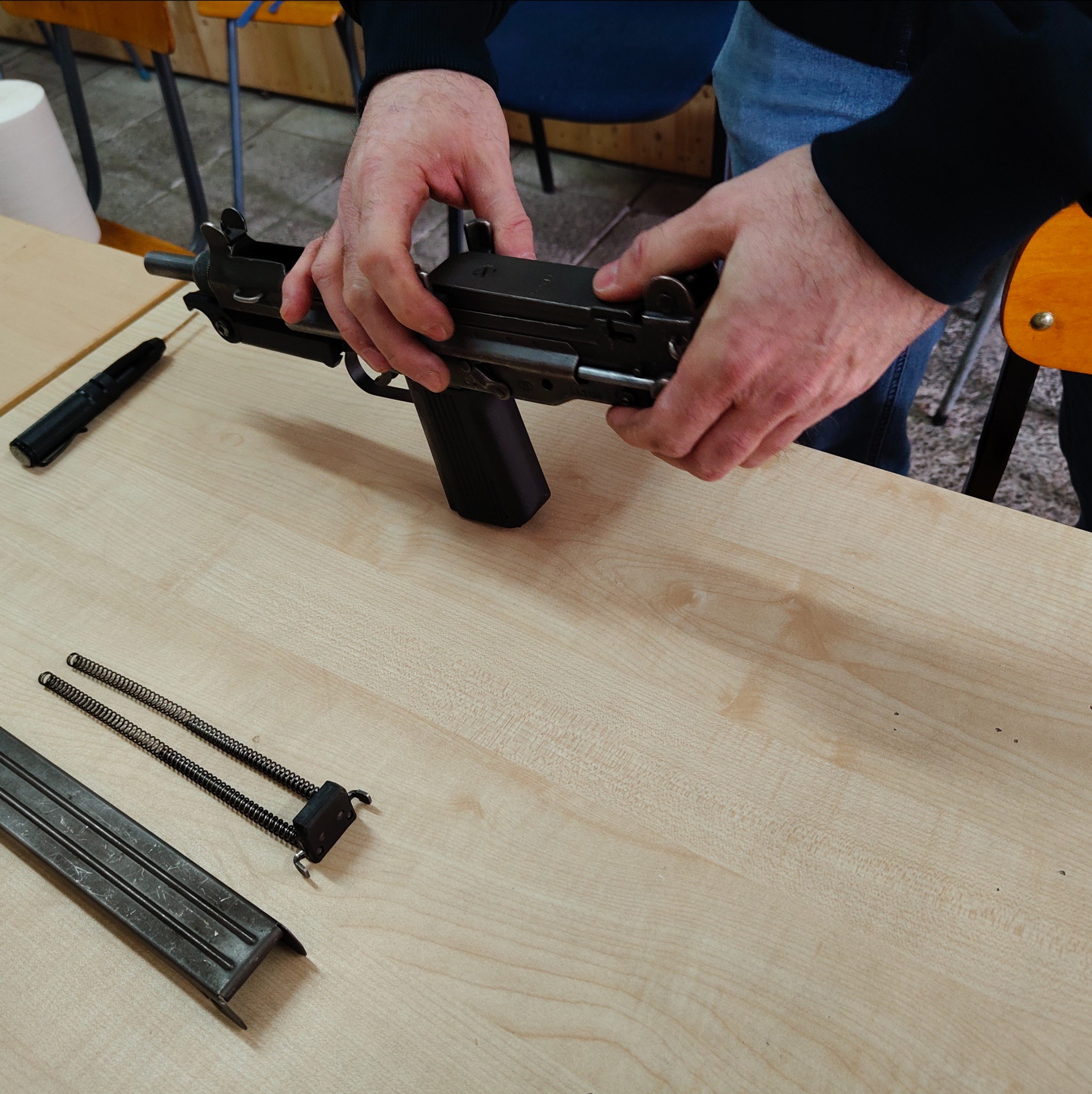 Rozkładanie broni palnej na drewnianym stole. Widoczne dłonie, magazynek, sprężyny i inne elementy. Szkolenie z zakresu obsługi broni.