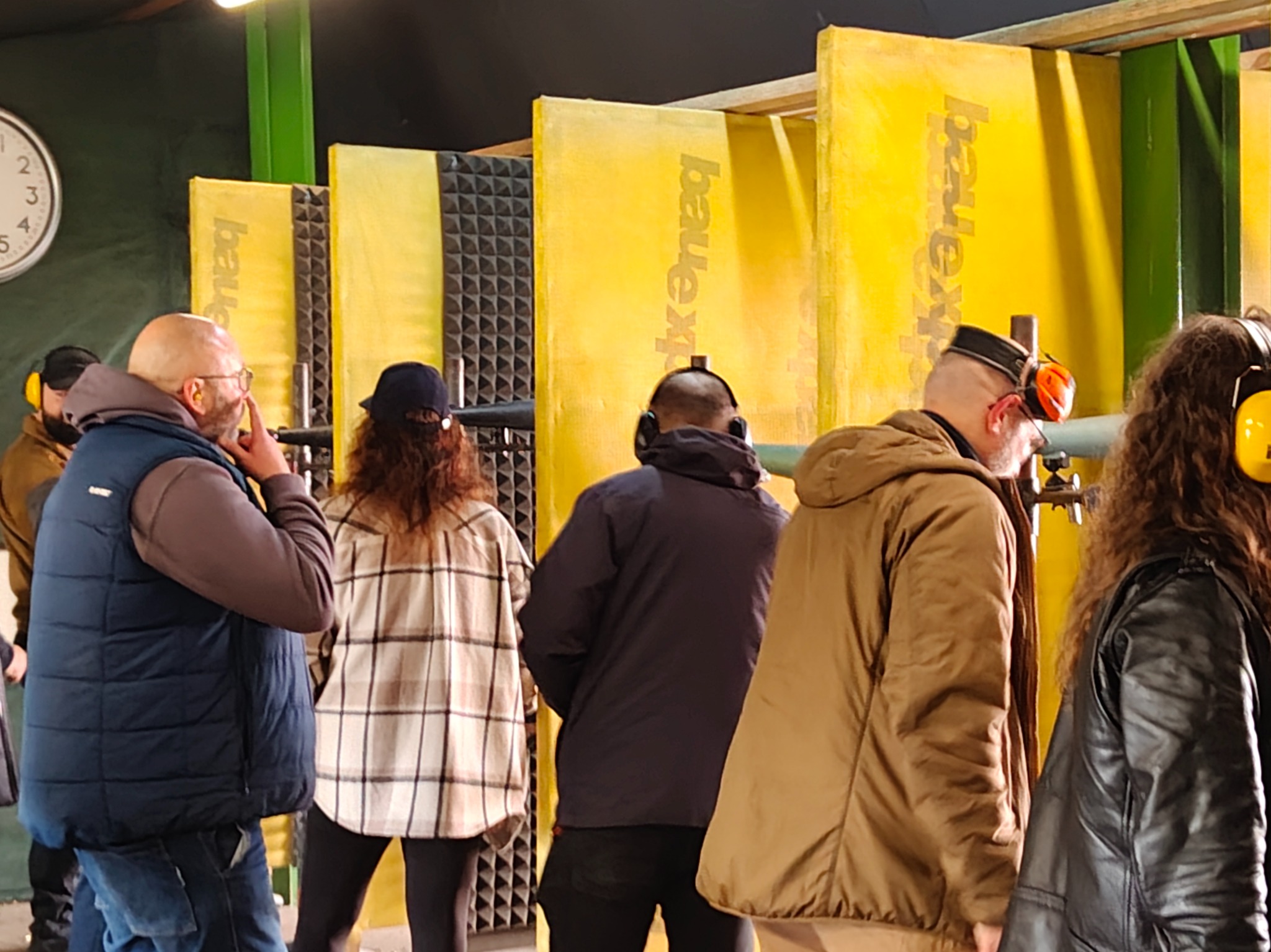 Grupa osób w słuchawkach na strzelnicy, żółte panele dźwiękochłonne w tle. Mężczyzna w okularach dotyka twarzy, pozostali odwróceni tyłem. Widoczny zegar na ścianie.