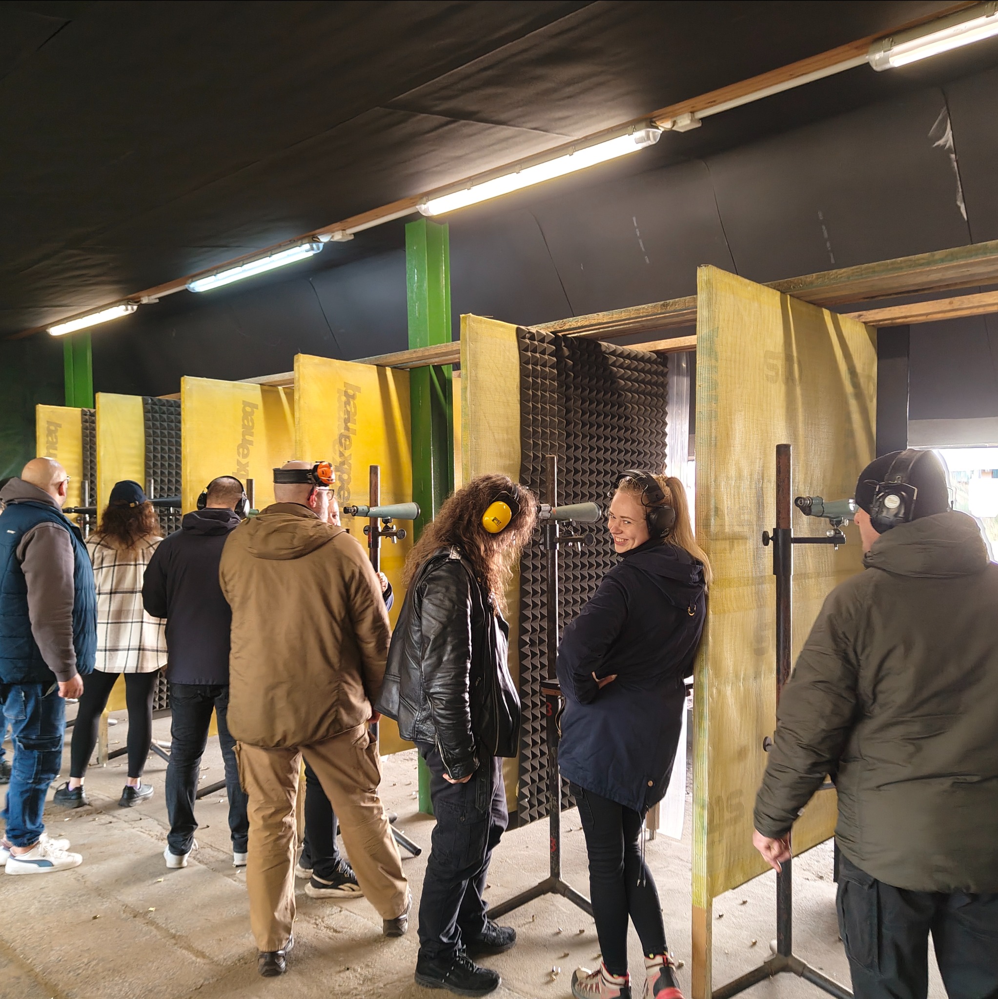 Grupa osób w słuchawkach na strzelnicy, przygotowujących się do strzelania. Widoczne stanowiska strzeleckie z osłonami akustycznymi i lunetami celowniczymi.