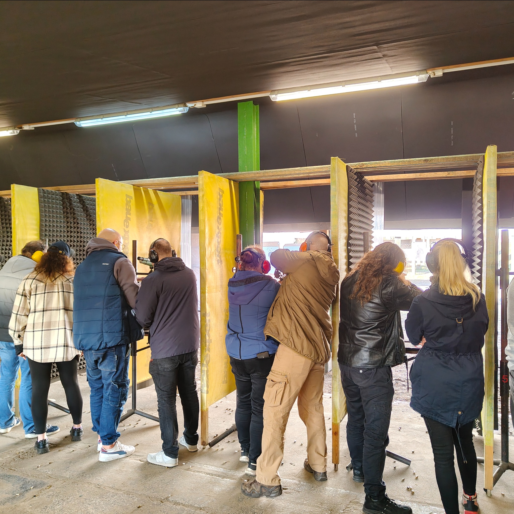 Grupa osób w słuchawkach ochronnych na strzelnicy, celuje do tarcz. Widoczne żółte przegrody i liczne łuski na podłodze. Uczestnicy w różnorodnych strojach.