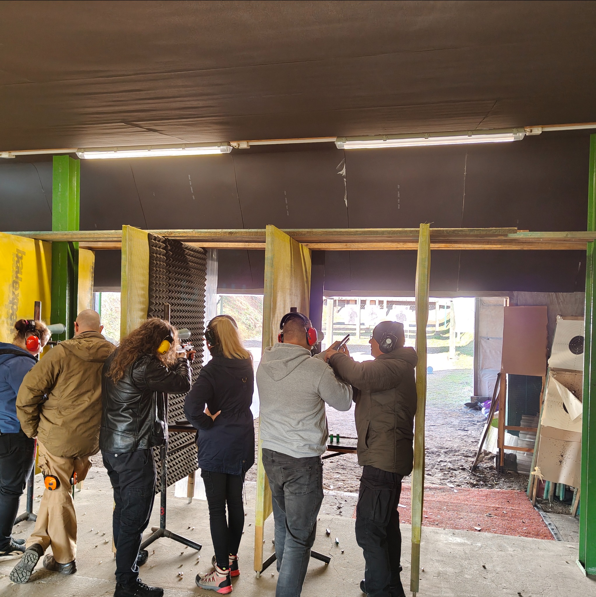 Grupa osób w słuchawkach na strzelnicy, celuje do tarcz. Widoczne stanowiska strzeleckie i ślady po pociskach. Kurs strzelecki w hali.