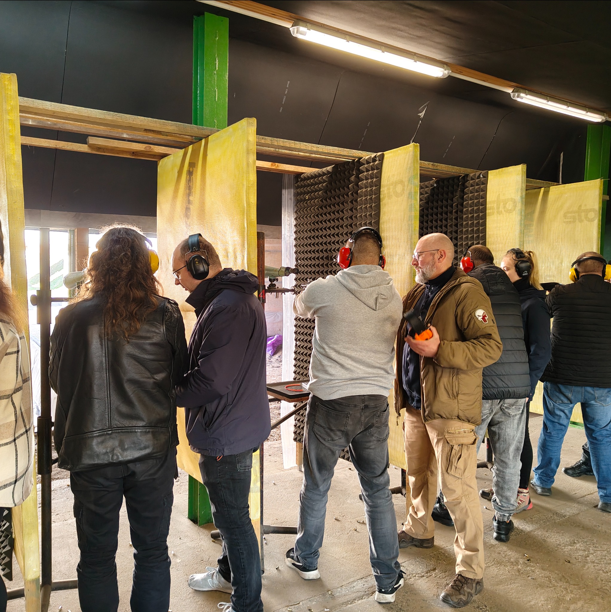 Grupa osób w słuchawkach na strzelnicy, jedna osoba celuje z pistoletu do tarczy. Widoczne stanowiska strzeleckie z żółtymi przegrodami i czarnym wygłuszeniem.