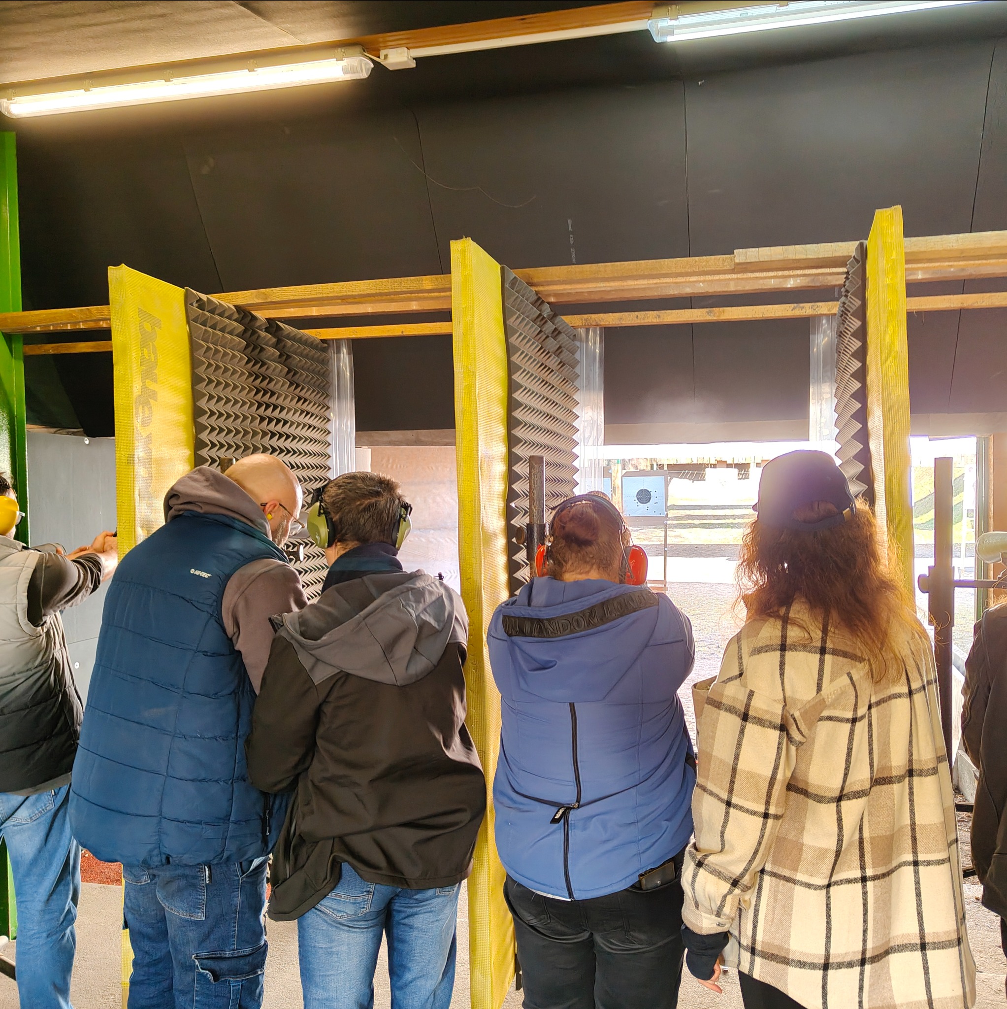 Grupa osób w słuchawkach ochronnych na strzelnicy, celuje do tarczy. Widoczne stanowiska z wygłuszeniem i oświetlenie. Uczestnicy kursu strzeleckiego.