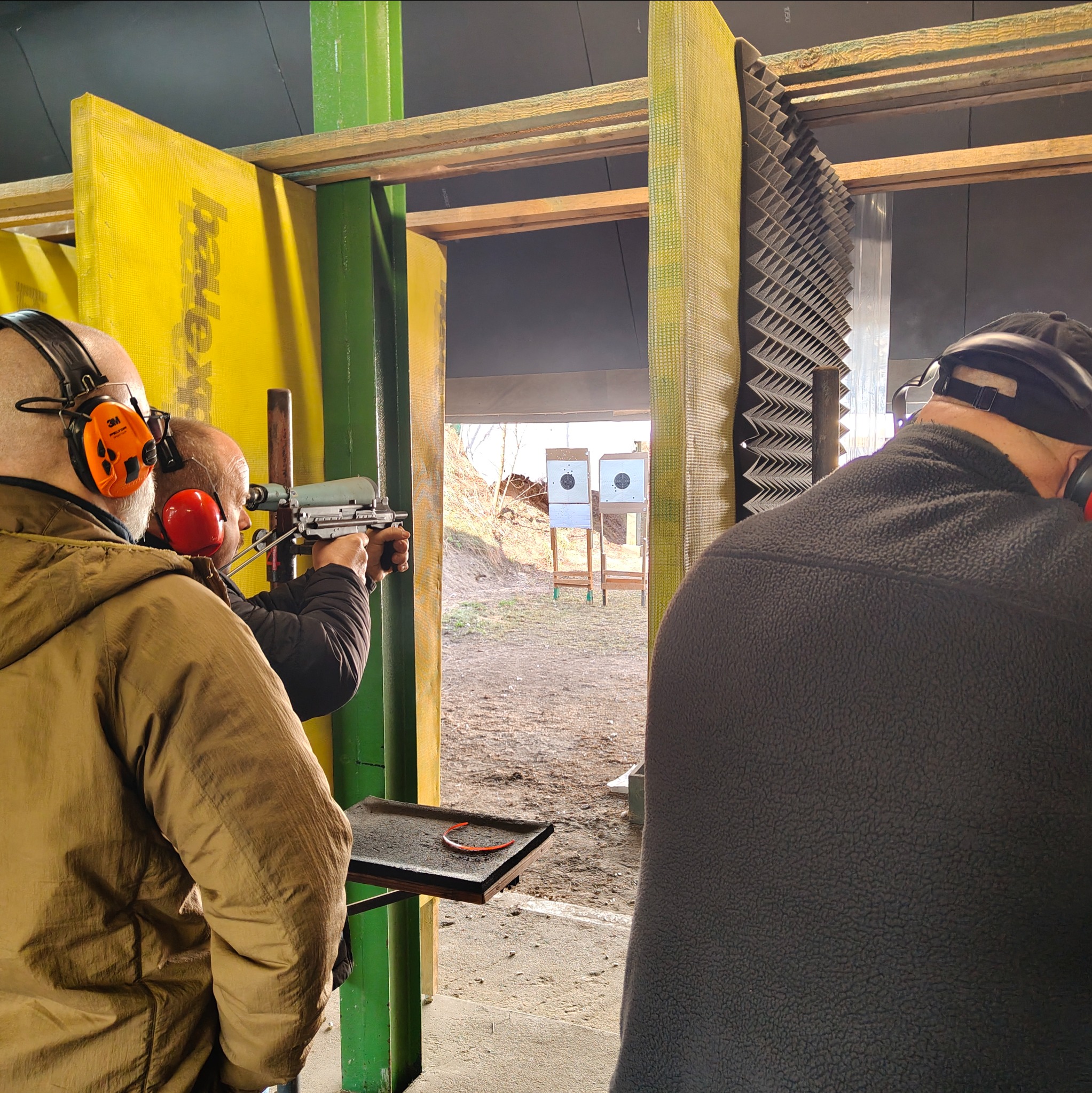 Strzelnica: mężczyzna w słuchawkach celuje z pistoletu do tarczy. Widoczne tarcze, osłony akustyczne i stanowiska strzeleckie. Uczestnicy kursu strzeleckiego.