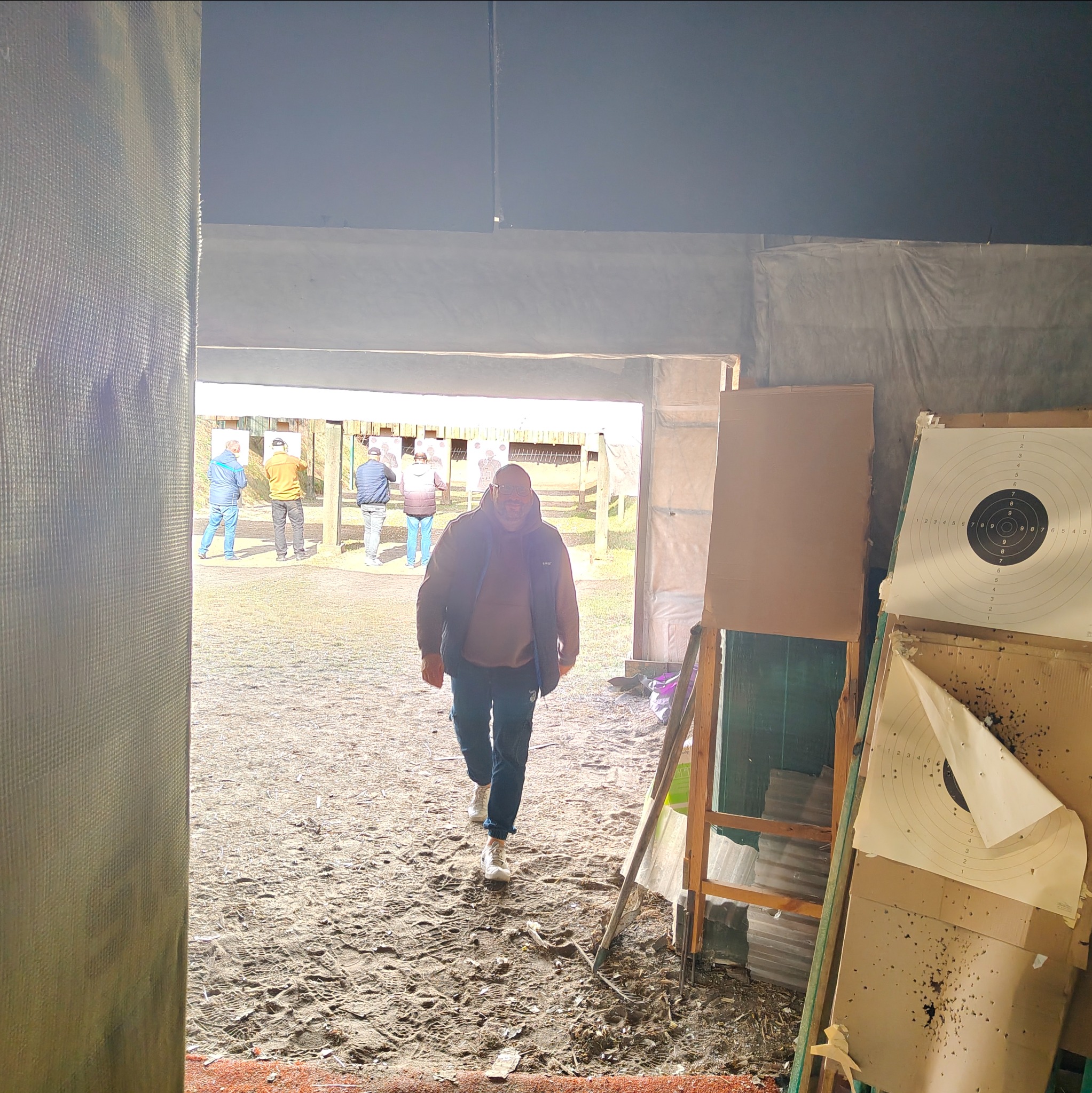Mężczyzna wychodzi z zadaszonej strzelnicy, w tle tarcze strzeleckie z widocznymi śladami po kulach. Grupa osób na stanowiskach strzeleckich w oddali.