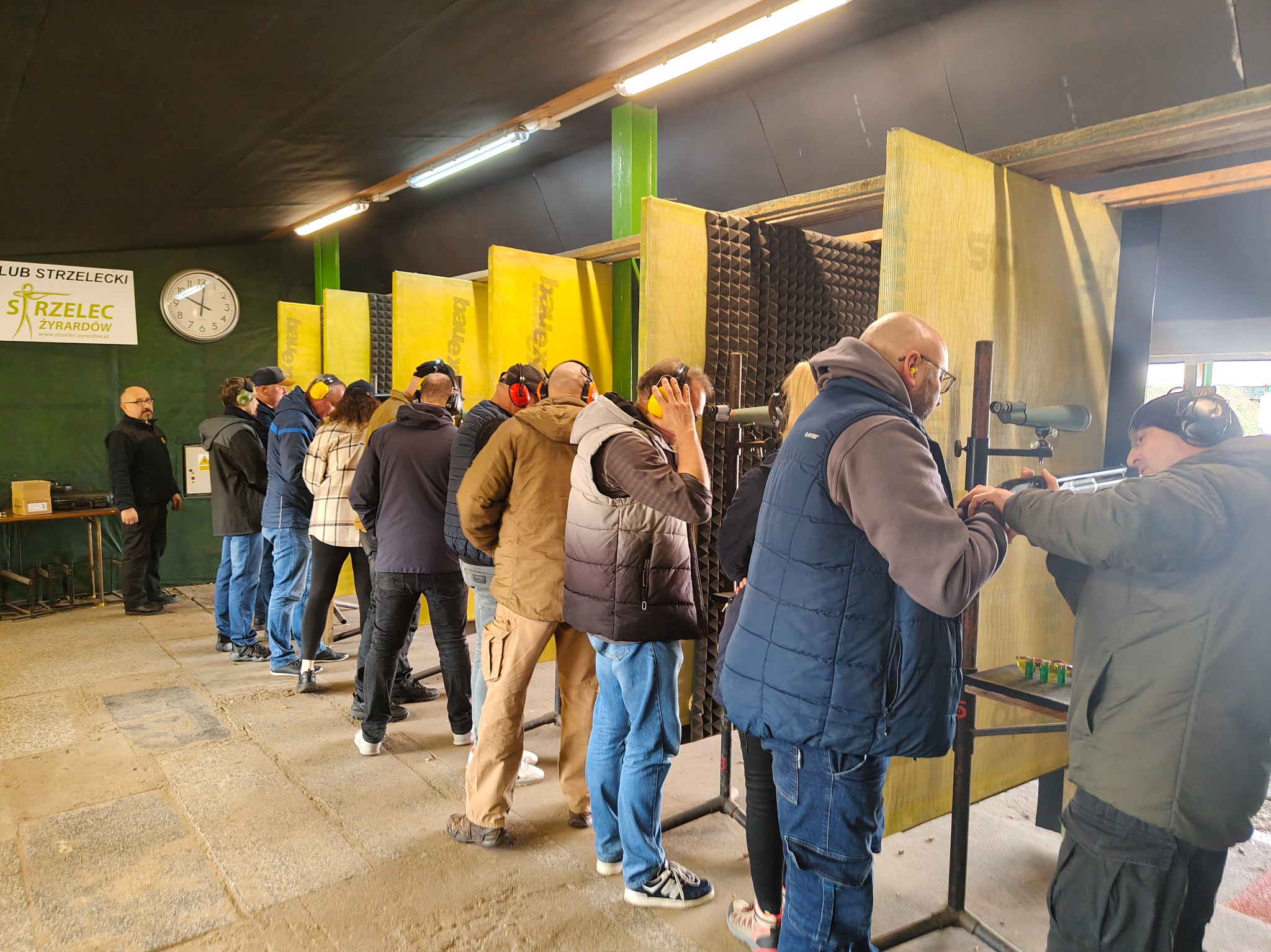 Grupa osób w słuchawkach na strzelnicy, przygotowujących się do strzelania. Widoczne tarcze, stanowiska strzeleckie i instruktorzy. Klub Strzelecki Żyrardów.