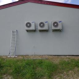 KLIMA-KURP - Trzy identyczne klimatyzatory Samsung zamontowane rzędem na jasnej ścianie budynku, z rozstawioną aluminiową drabiną po lewej stronie, widok z poziomu gruntu na zarośnięty trawnikiem teren przed...