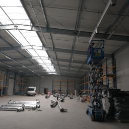 KLIMA-KURP - Montaż systemu wentylacji w hali: pracownik na podnośniku koszowym instaluje rury wentylacyjne pod sufitem, w tle składowane elementy systemu i biały bus.