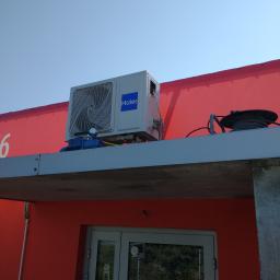 KLIMA-KURP - Zewnętrzna jednostka klimatyzacji Haier zamontowana na płaskim dachu budynku o czerwonej fasadzie, widoczne narzędzia serwisowe i przewody, na drzwiach wejściowych naklejka informująca o obowiązku...