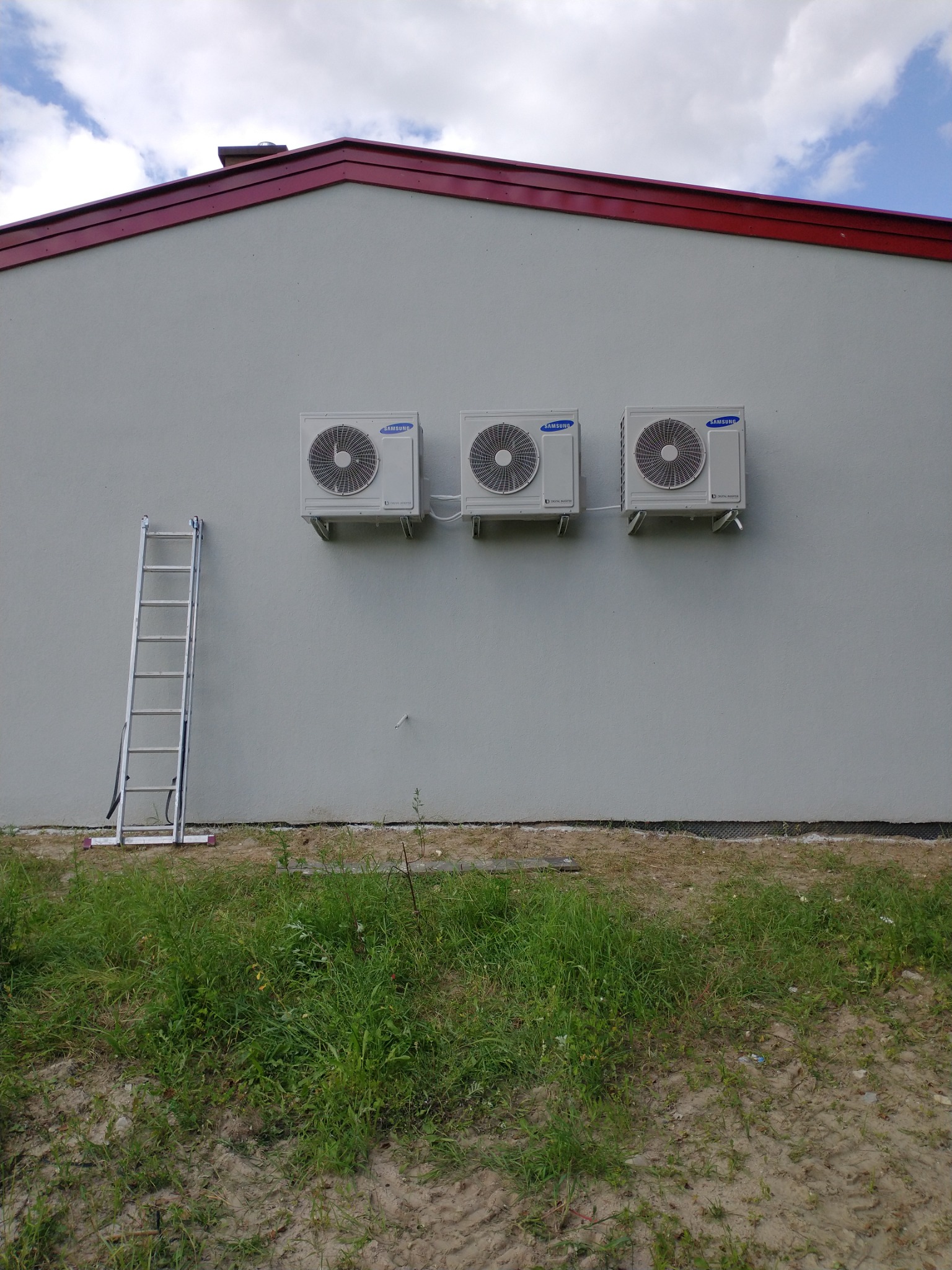 Trzy identyczne klimatyzatory Samsung zamontowane rzędem na jasnej ścianie budynku, z rozstawioną aluminiową drabiną po lewej stronie, widok z poziomu gruntu na zarośnięty trawnikiem teren przed...
