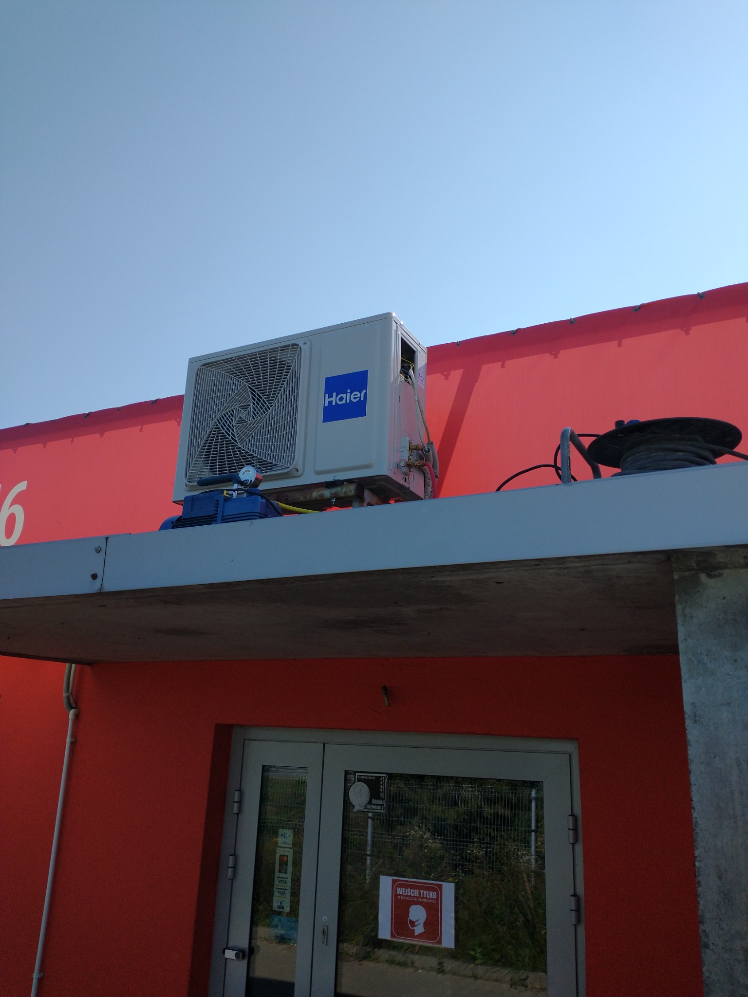 Zewnętrzna jednostka klimatyzacji Haier zamontowana na płaskim dachu budynku o czerwonej fasadzie, widoczne narzędzia serwisowe i przewody, na drzwiach wejściowych naklejka informująca o obowiązku...