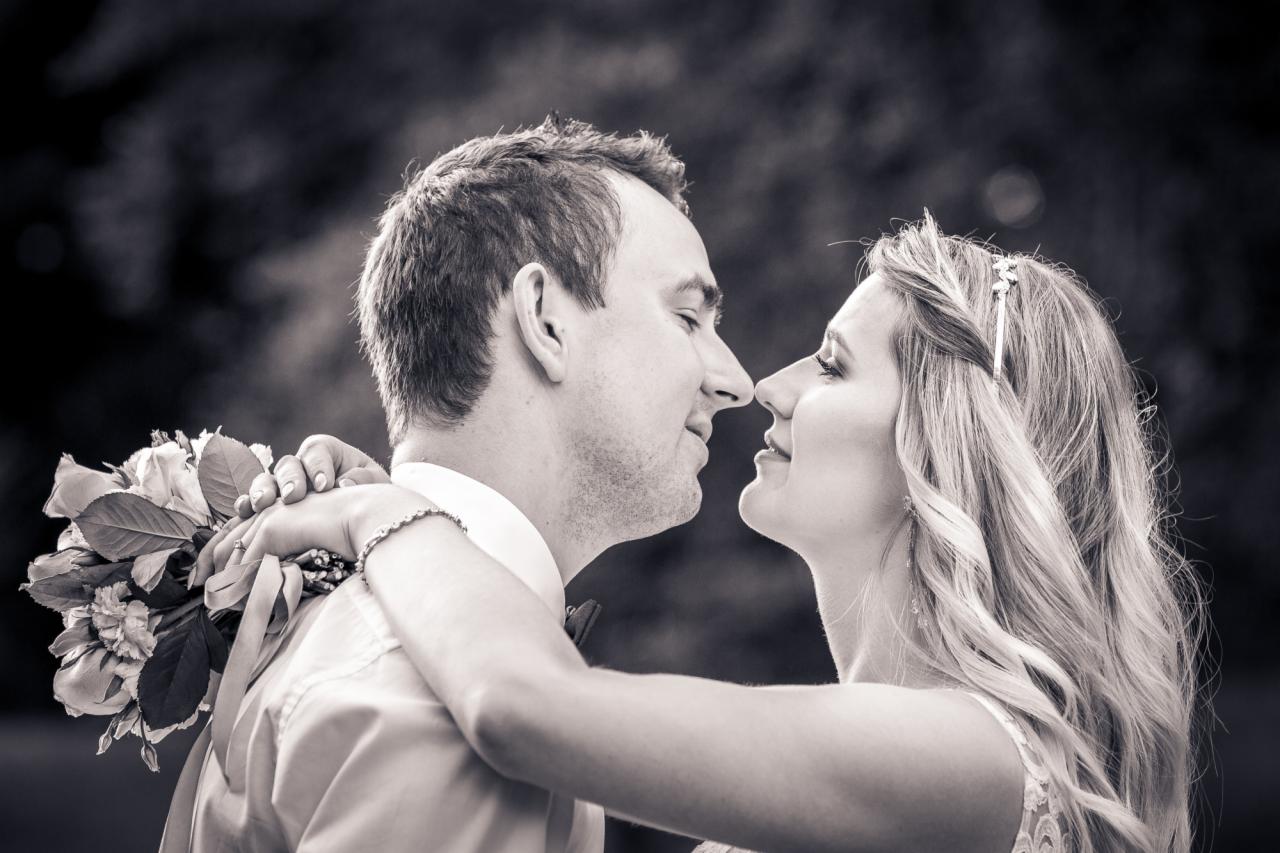 Czarno-białe zbliżenie pary młodej, nosy prawie się stykają, panna młoda z kwiatami opiera ręce na ramionach pana młodego, delikatny wianek we włosach.