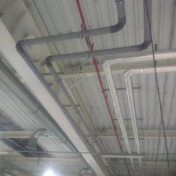 Alsamet Jakub Czabanowski - Różne rodzaje rur instalacyjnych (szare, białe, metalowe) zamontowane pod sufitem hali przemysłowej, widoczne mocowania i elementy konstrukcyjne.