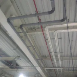 Różne rodzaje rur instalacyjnych (szare, białe, metalowe) zamontowane pod sufitem hali przemysłowej, widoczne mocowania i elementy konstrukcyjne.