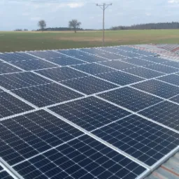 Rząd paneli słonecznych na blaszanym dachu budynku, z polami i drzewami w tle pod pochmurnym niebem.