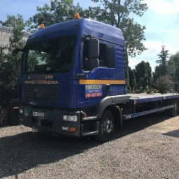 Niebieska laweta MAN TGL 12.240 z napisem 'Pomoc Drogowa' oraz numerem telefonu, zaparkowana na żwirowym podłożu przed budynkiem.