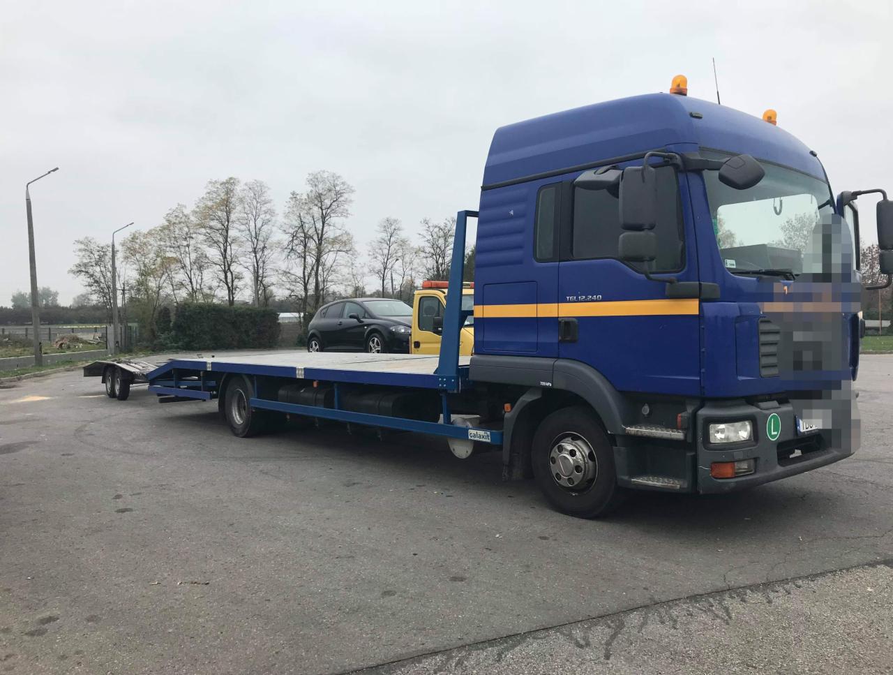 Niebieska laweta MAN TGL 12.240 z żółtym pasem, zaparkowana na szarym asfalcie; na platformie widoczny czarny samochód osobowy.