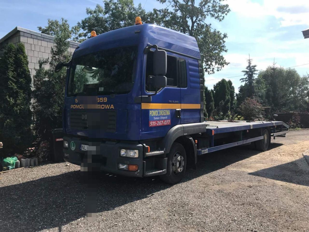 Niebieska laweta MAN TGL 12.240 z napisem 'Pomoc Drogowa' oraz numerem telefonu, zaparkowana na żwirowym podłożu przed budynkiem.