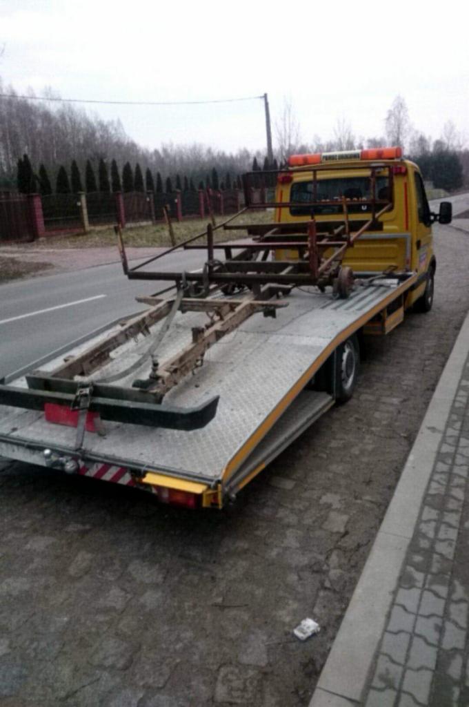 Żółta laweta z elementami karoserii przewożonymi na platformie, zaparkowana na poboczu drogi w pochmurny dzień.