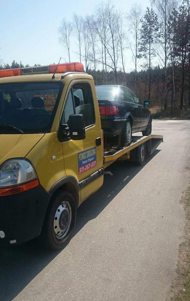 Żółta laweta z czarnym samochodem osobowym marki Audi na platformie, widok z boku na drodze asfaltowej, w tle drzewa bez liści.