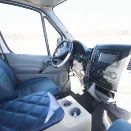 Wnętrze busa z niebieską tapicerką w romby na siedzeniach i jasnym beżowym podłokietnikiem z uchwytami na napoje, widok z perspektywy pasażera.