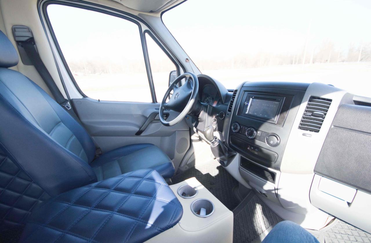 Wnętrze busa z niebieską tapicerką w romby na siedzeniach i jasnym beżowym podłokietnikiem z uchwytami na napoje, widok z perspektywy pasażera.