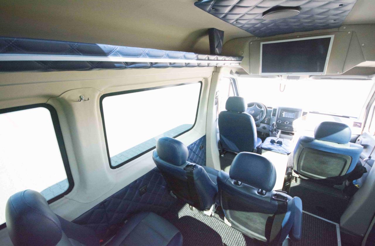 Wnętrze luksusowo wykończonego busa z pikowaną tapicerką w odcieniach beżu i granatu, z zamontowanym telewizorem i dodatkową półką nad oknami.