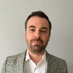 Portret mężczyzny z lekkim zarostem, ubranego w białą koszulę i szarą marynarkę w kratę, na jasnoszarym tle.