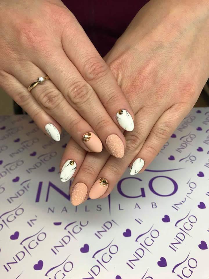 Dłonie kobiety z manicure hybrydowym: dwa paznokcie beżowe z brokatem i złotymi zdobieniami, trzy białe z marmurkowym wzorem i czarną linią oraz złotym akcentem, pierścionek na palcu serdecznym...