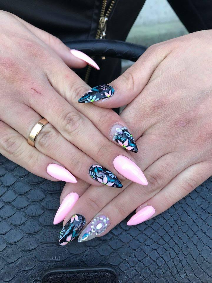 Dłonie z pomalowanymi paznokciami w kształcie szpiców: trzy w kolorze jasnego różu, dwa z ciemnym tłem i kwiatowym wzorem, jeden z przezroczystą bazą i zdobieniami z cyrkonii.