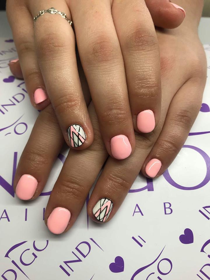 Dłonie z pomalowanymi na jasny różowo paznokciami, jeden paznokieć z geometrycznym wzorem w czerni i bieli, na tle z logo Indigo.