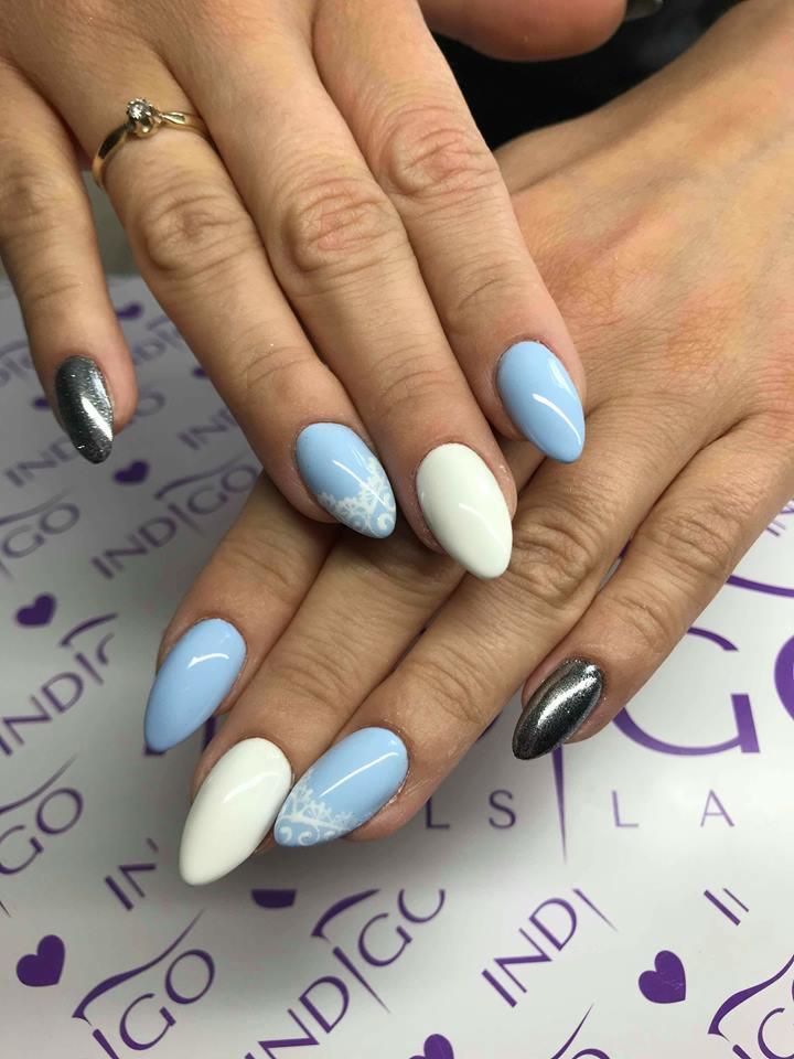 Dłonie z pomalowanymi paznokciami w kolorach: błękitny z białym wzorem śnieżynki, biały i srebrny brokat. Na palcu serdecznym złoty pierścionek z oczkiem. Tło stanowi firmowy podkład z logo.