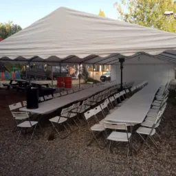 Biały namiot imprezowy z rozstawionymi stołami nakrytymi białymi obrusami i białymi krzesłami, na żwirowym podłożu, w tle plac zabaw i pojazd wojskowy.