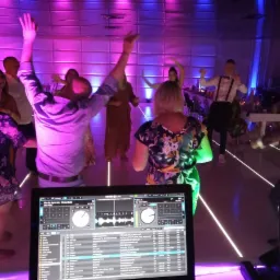 Parkiet taneczny wypełniony rozbawionymi gośćmi, oświetlony dynamicznymi światłami, z widocznym w pierwszym planie ekranem miksera DJ-a, grającego muzykę.