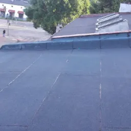 Płaski, ciemny dach pokryty papą termozgrzewalną z widocznymi łączeniami, w tle budynki mieszkalne i ulica z przechodniem.