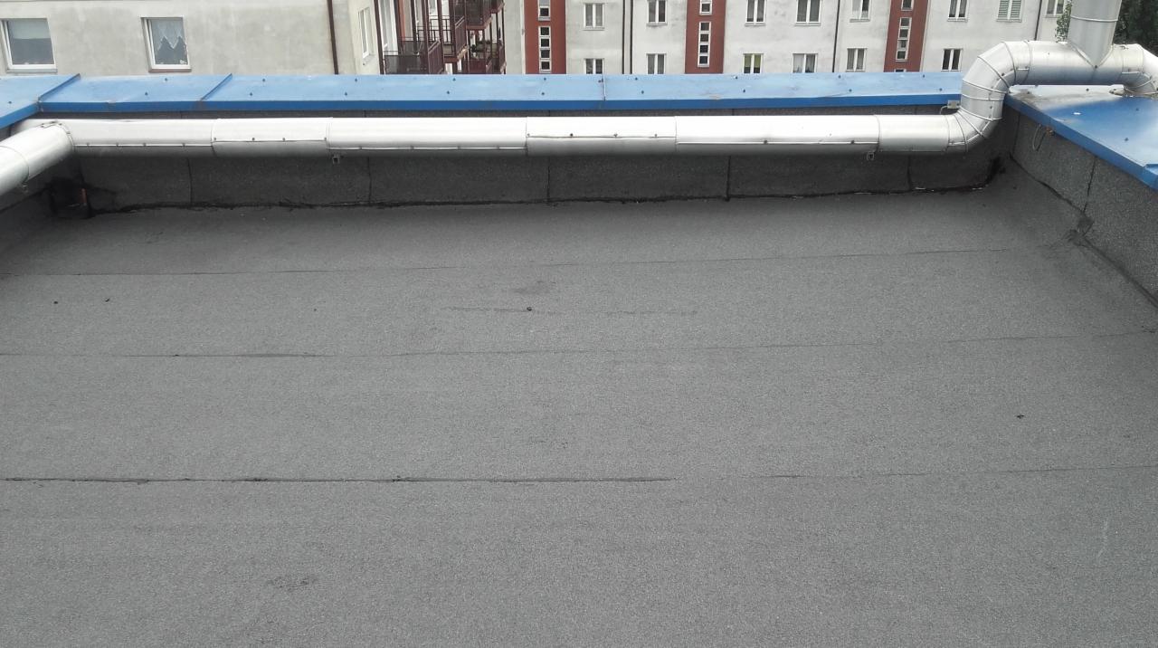 Płaski dach pokryty papą termozgrzewalną z widocznymi srebrnymi rurami wentylacyjnymi i niebieskimi elementami obróbek blacharskich na tle budynków mieszkalnych.