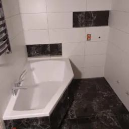 Nowoczesna łazienka z białą wanną obudowaną czarnym marmurem, białe płytki ścienne z poziomymi wstawkami z czarnego marmuru, podłoga wyłożona czarnymi płytkami imitującymi marmur, widoczne elementy...