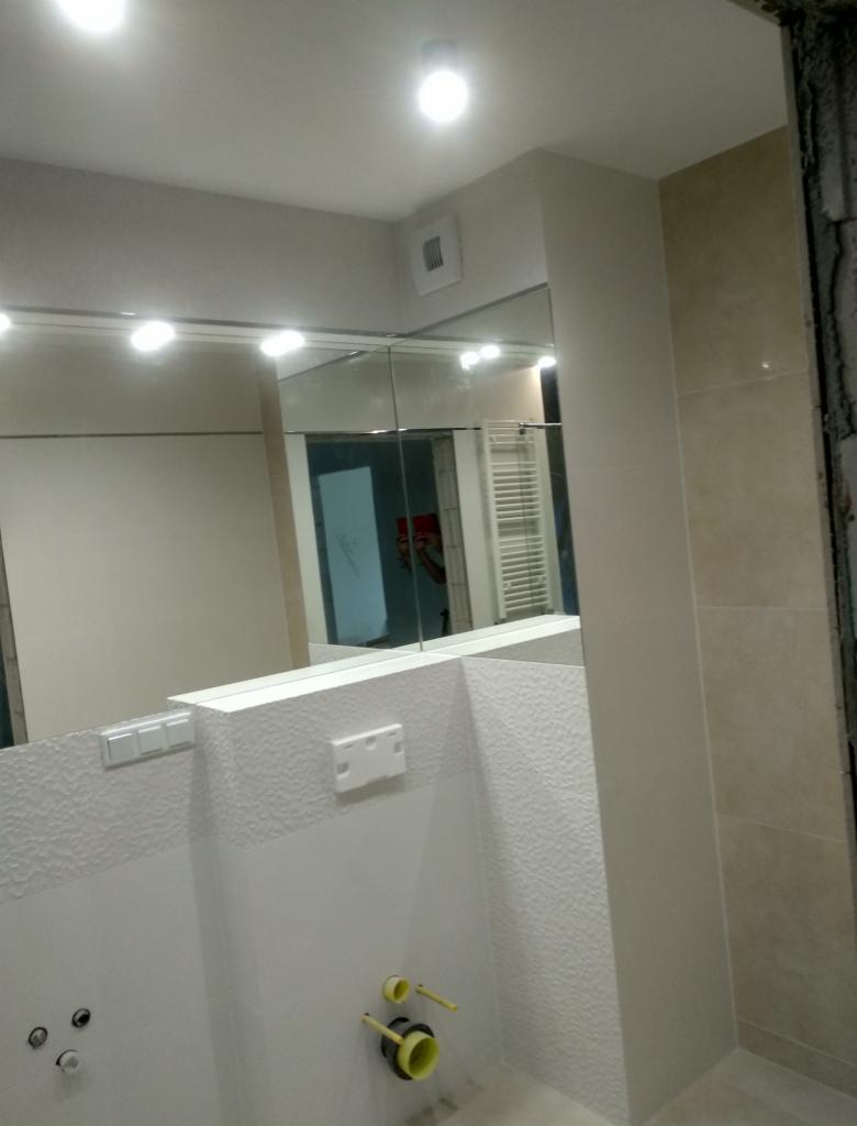 Nowoczesna łazienka w trakcie wykańczania: widoczne fragmenty lustra z oświetleniem, białe ściany z teksturą, instalacja pod umywalkę z żółtymi elementami, płytki w odcieniach beżu na ścianie po...