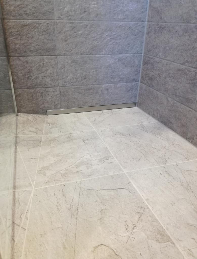 Wykończony odpływ liniowy w kabinie prysznicowej wyłożonej szarymi płytkami imitującymi kamień.