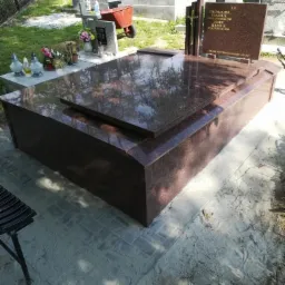 Nowoczesny nagrobek z ciemnego, polerowanego granitu z krzyżem i tablicą inskrypcyjną, otoczony innymi grobami na cmentarzu w słoneczny dzień.