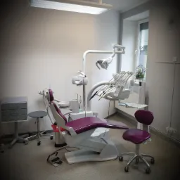 Nowoczesny gabinet dentystyczny z fotelem w kolorze fuksji, oświetleniem sufitowym i narzędziami na białym ramieniu. W tle okno z dekoracyjną klatką dla ptaków i zegar na ścianie.