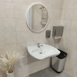 Umywalka z baterią, okrągłe lustro, dozownik mydła i podajnik ręczników w jasnej toalecie wyłożonej beżowymi płytkami, z dekoracyjnym bukietem w wazonie.