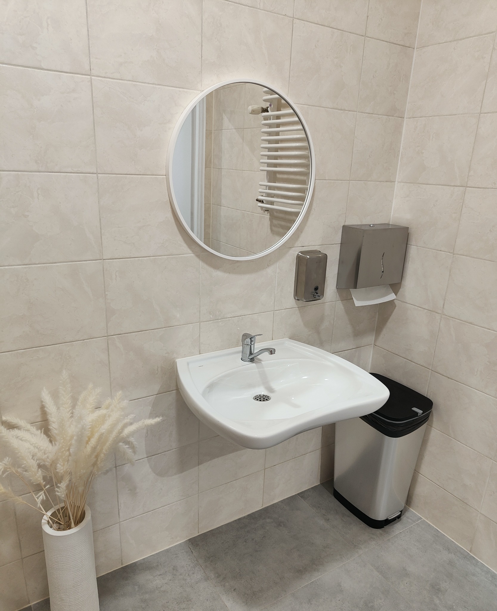 Umywalka z baterią, okrągłe lustro, dozownik mydła i podajnik ręczników w jasnej toalecie wyłożonej beżowymi płytkami, z dekoracyjnym bukietem w wazonie.