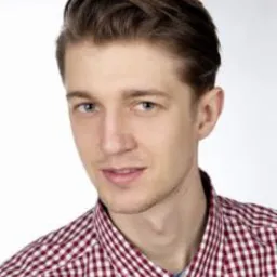 Portret młodego mężczyzny w koszuli w czerwoną kratę, z delikatnym uśmiechem, na jasnym, neutralnym tle.