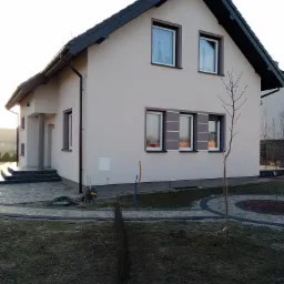Nowo wybudowany, otynkowany dom jednorodzinny z grafitową dachówką, widok z ogrodu przed budynkiem. Zauważalna świeżo położona kostka brukowa i przygotowane podłoże pod trawnik.