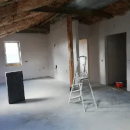 Poddasze w trakcie wykańczania: ściany pokryte świeżym, jasnym tynkiem, widoczne instalacje elektryczne, drabina malarska, fragment izolacji dachu i element konstrukcyjny z drewna.