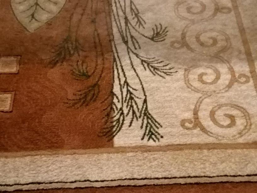 Fragment brązowo-beżowego dywanu z wzorem liści i spiralnych ornamentów, widoczne delikatne cienie na powierzchni.