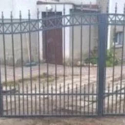 Metalowa brama wjazdowa i furtka z dekoracyjnymi elementami w kształcie krzyżyków, pomalowana na ciemny kolor, na tle budynku mieszkalnego.