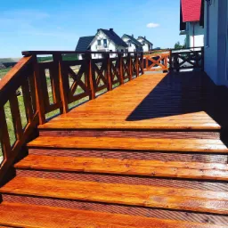 Nowoczesny, lakierowany taras z drewna w Pelplinie, z balustradą w kształcie krzyżyków, prowadzący do domu z czerwonym dachem na tle błękitnego nieba.